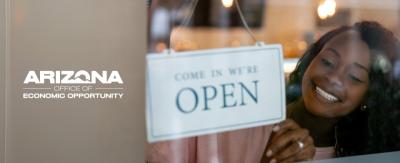 Microbusiness owner putting up sign saying "Come in. We're Open." with the Arizona Office of Economic Opportunity logo on the side